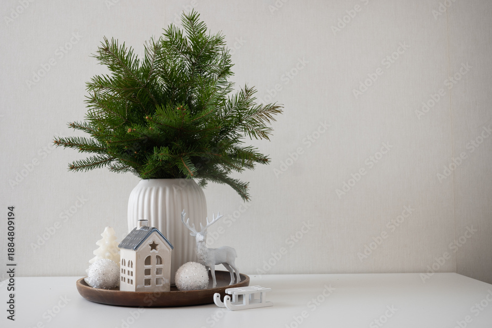 Fototapeta premium Minimal winter home decor with fir branches, ceramic house and deer figurine arranged on white table against neutral background.
