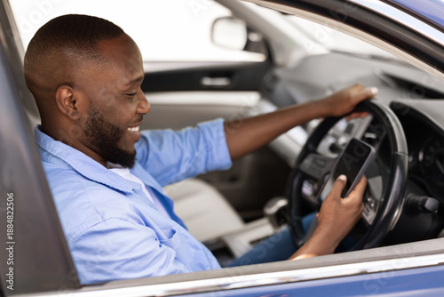 Foto Smiling black guy using smartphone, reading and answering messages or checking emails while driving car