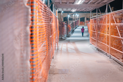 Night pedestrian corridor at Ljubljana train station renovation: temporary walkway lined with orange safety fencing and scaffolding, lights overhead, a lone passerby moving through the construction