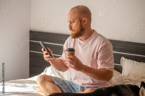 A bald man with a beard in a pink T-shirt drinks morning coffee and looks at a smartphone while sitting on a bed in a bedroom