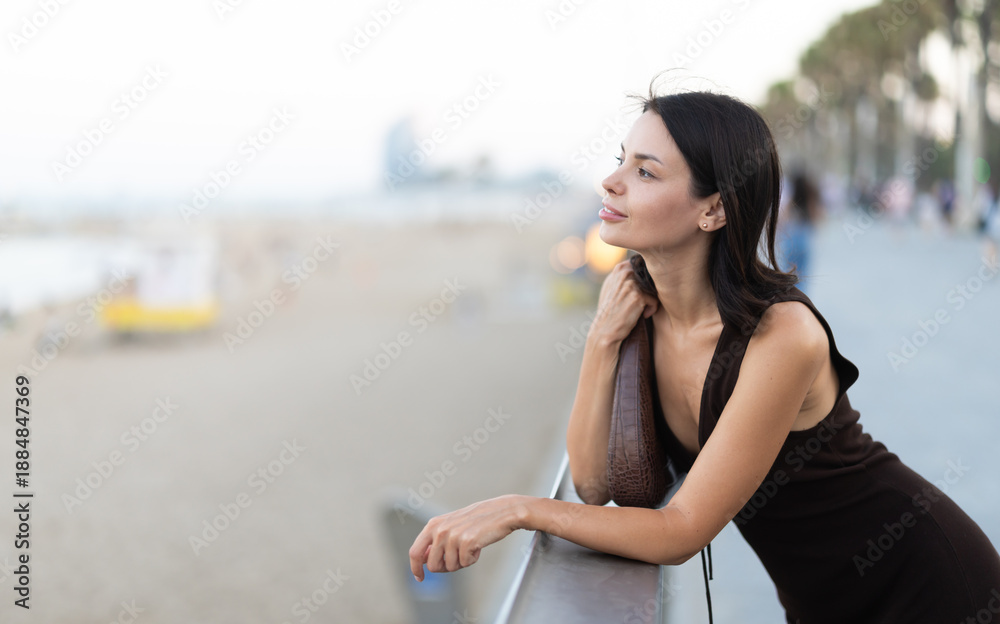 Fototapeta premium Elegant young woman in summer dress leaning on parapet at Barceloneta beach promenade