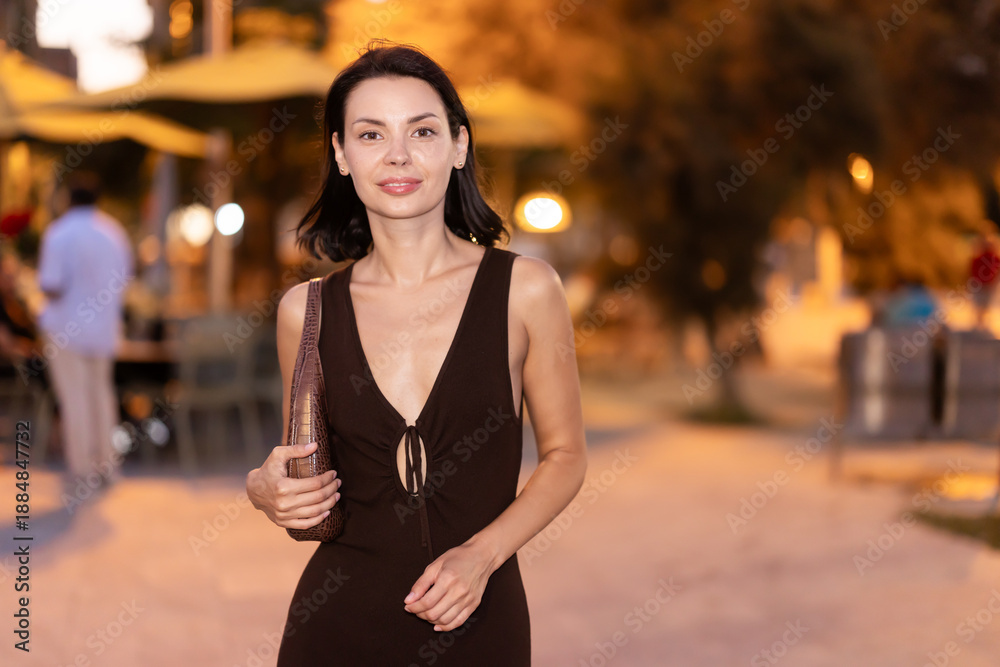 Fototapeta premium Smiling young woman walking through urban street with lively atmosphere under evening lights in Barcelona