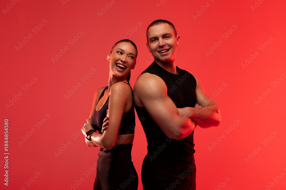 Obraz premium Happy sporty couple standing back to back and smiling, man and woman wearing black workout gear, posing over red neon studio background