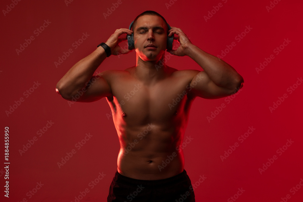Fototapeta premium Male athlete with bare torso listening to music in wireless headphones with closed eyes, standing over red studio background. Sport and technology concept