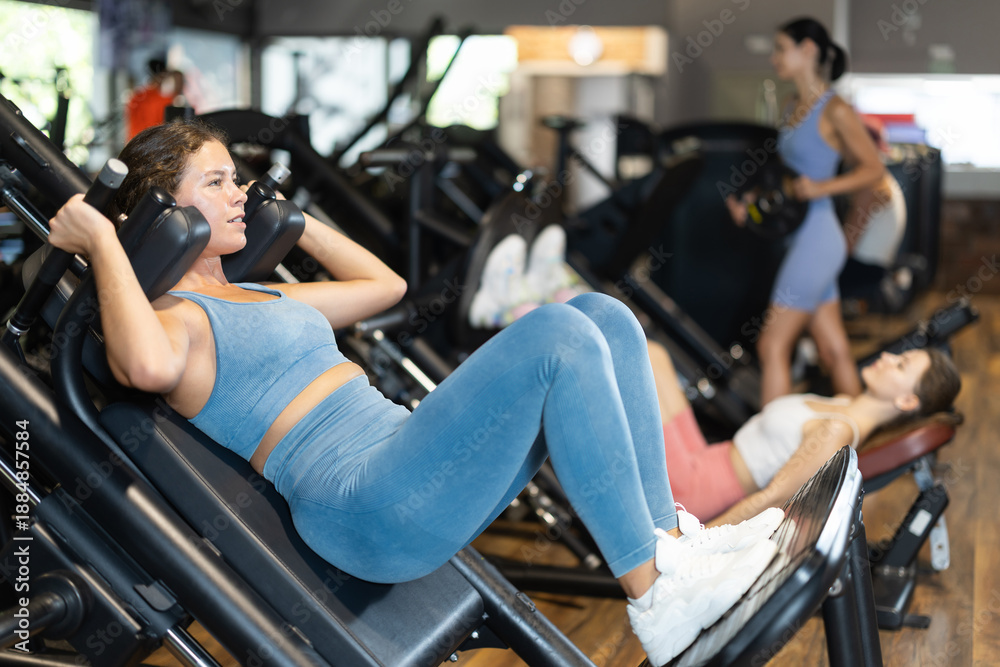 Obraz premium Athletic woman in top and leggings doing exercises in hack squat machine. Young woman training glutes in the gym