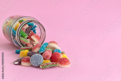 Front view of colorful sugared gummy candies spilling out of an overturned glass jar on the left side of a bright pink background with copy space on the right. Close-up, shallow depth of field