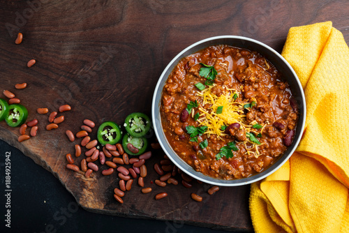 Φωτογραφία A rustic overhead view of homemade beef chili served in bowls and topped with shredded cheddar cheese, fresh herbs, and sliced jalapeños