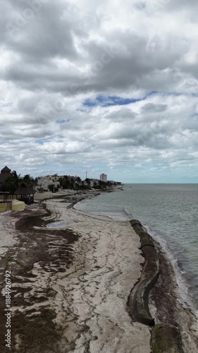Aerial drone flying forward along the shoreline in Puerto Progreso, revealing a quiet beach, coastal buildings, and calm sea under an overcast sky, captured in D-Log.