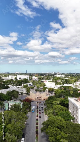 Vertical aerial view in Mérida, Yucatán, as the drone flies forward over Paseo Montejo toward the Altar a la Patria roundabout, with lush trees, city traffic, and a blue, cloud-filled sky.