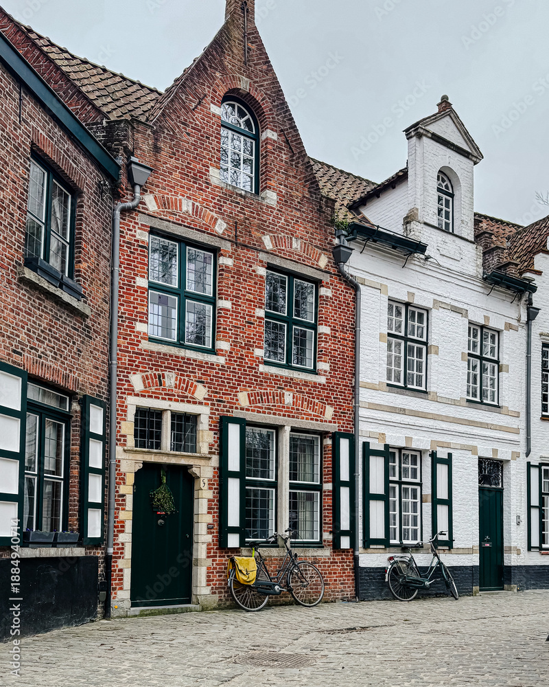 Naklejka premium Bruges, Belgium – December 27, 2025. Traditional brick houses with white facades, balconies and green shutters on a quiet cobblestone street in the historic old town.