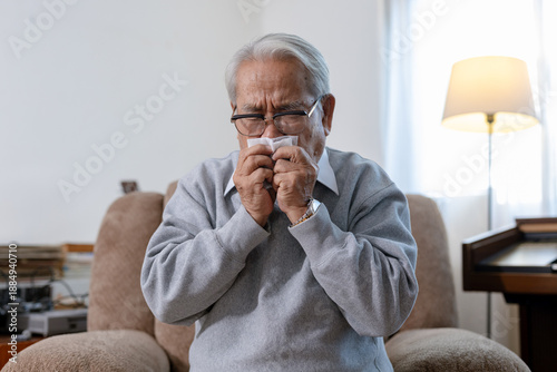 Tableau sur toile Asian senior man sitting on sofa and sneezing into tissue at home, suffering from cold, flu, seasonal allergy