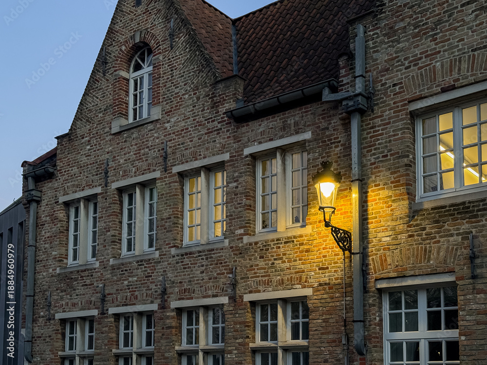 Naklejka premium Bruges, Belgium – December 27, 2025. Historic brick house facade with traditional Flemish gable, warm street lantern and illuminated windows at blue hour, atmospheric European old town scene.
