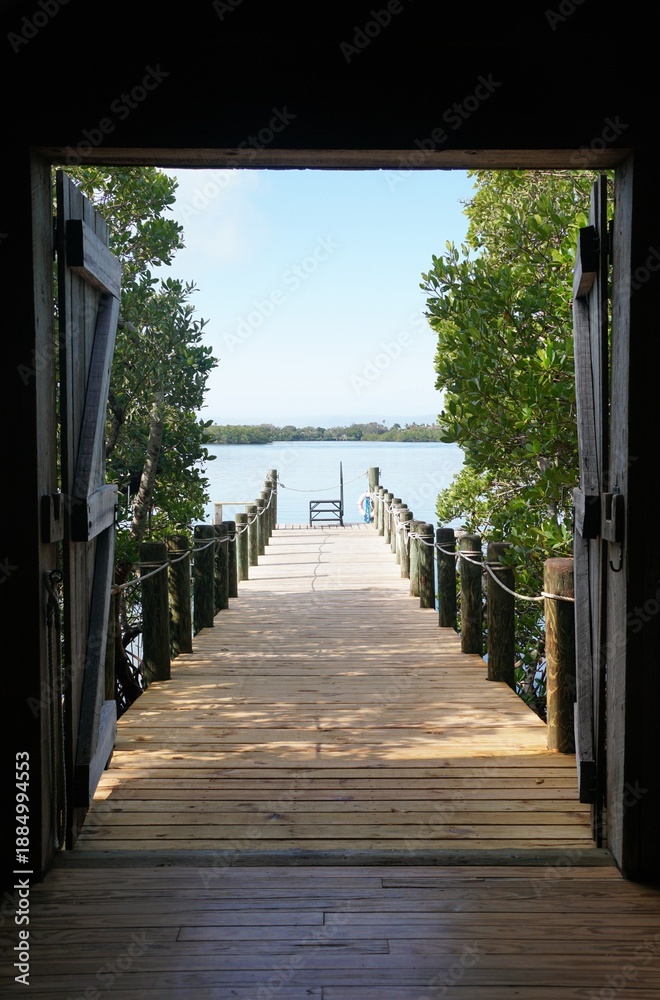 Fototapeta premium A wooden dock extends toward calm water, framed by open barn-style doors and lush green mangroves near Mary Selby Gardens Historic Spanish Point, Sarasota, Florida, U.S