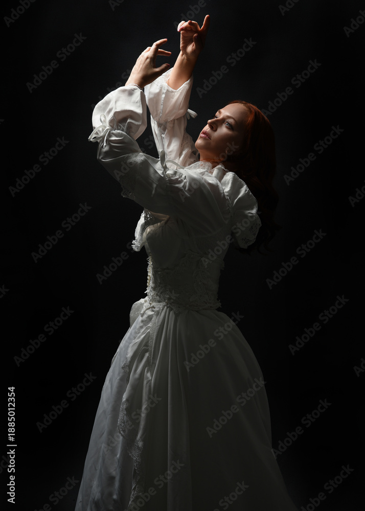 Fototapeta premium close up portrait beautiful female model wearing antique white historical ghost bride wedding dress gown. Overhead silhouette lighting, dark studio background. looking up towards light, hands reaching