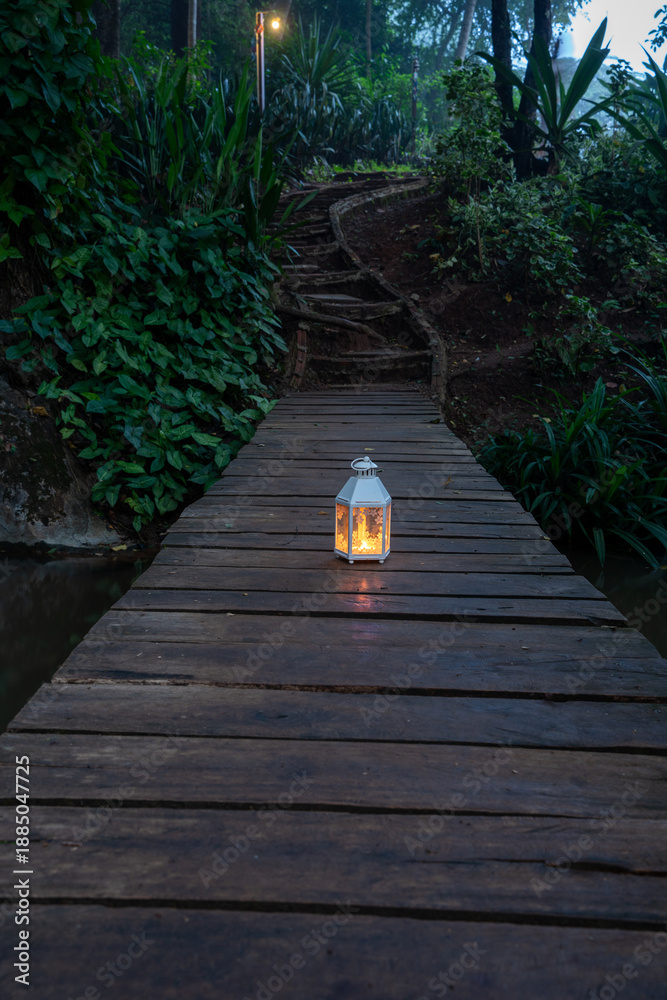 Fototapeta premium A glowing white vintage lantern placed on a wooden boardwalk path in a lush tropical garden at twilight. Serene outdoor scene with warm light and dense jungle foliage background.