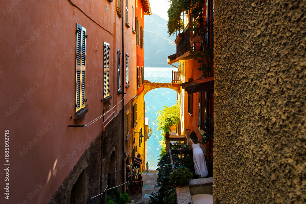Obraz premium Locals enjoy the water views from a narrow colorful alley with an archway leading to the shores of Lake Como in the lakefront resort village of Varenna.