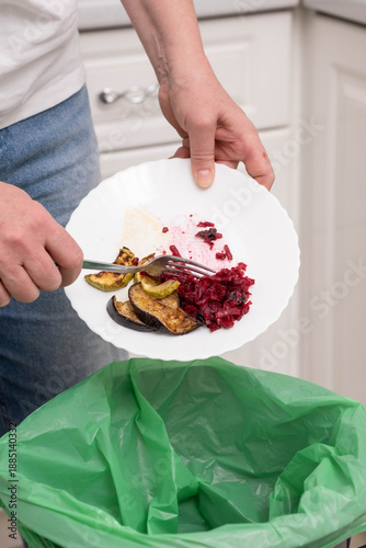 Fotografie A person is discarding leftover food, roasted vegetables and beetroot from a white plate into a trash bin with a green liner