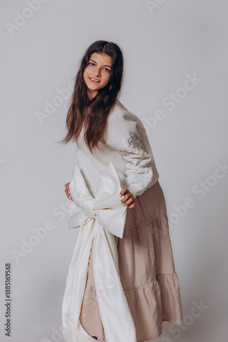The model is a young beautiful brunette girl with a big white bow in a white sweater and skirt in the studio