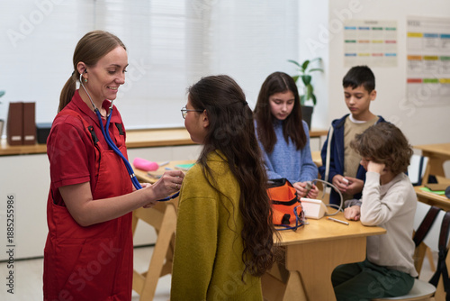 Caucasian young adult woman using stethoscope examining Asian preteen girl, while diverse group of children sitting at table engaging with medical equipment in classroom setting