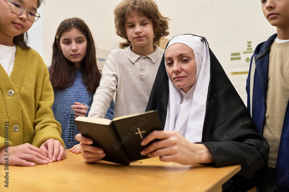 Fototapeta premium Caucasian middle aged nun sitting at table reading Bible while multiethnic group of children and teenagers standing around listening attentively in classroom setting