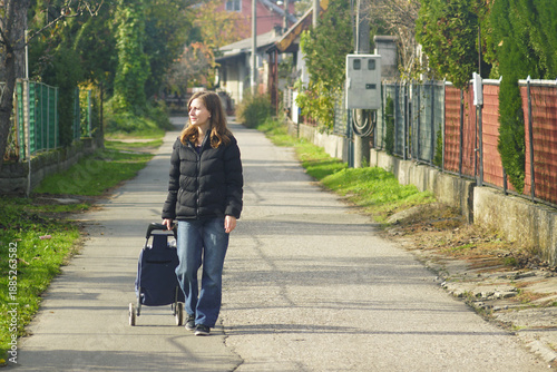Smiling young woman walks with a shopping cart along a sunlit, empty residential street. Represents independent daily mobility, low-traffic neighborhood living, and post-peak consumption lifestyle.