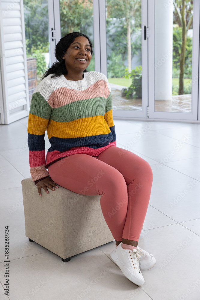 Obraz premium African American woman sitting on beige ottoman by glass doors, wearing striped knit, smiling up