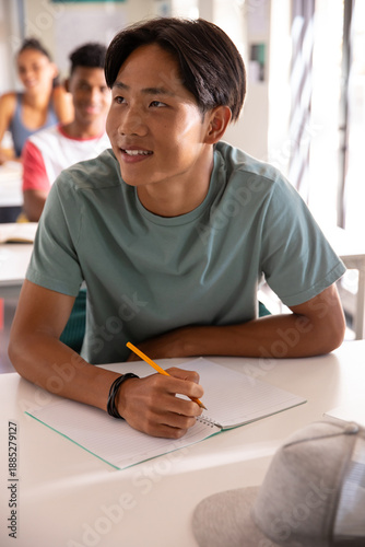 Diverse teenage classmates sitting at desks in classroom, writing in notebooks with pencil