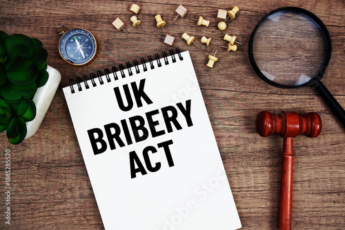 UK Bribery Act (UKBA) message written on notepad with wood gavel and magnifying glass flat lay on wooden background