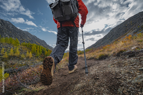 Low angle view of a person hiking up a rocky mountain trail during autumn, wearing a red jacket, black trousers, and using a trekking pole.