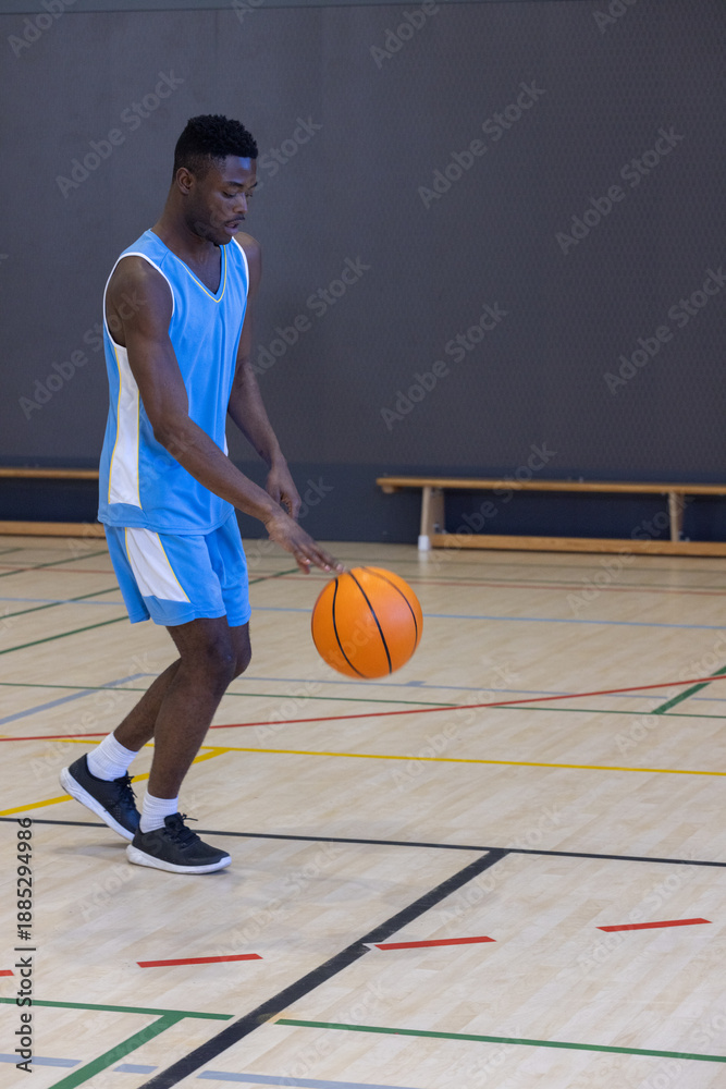 Fototapeta premium African american man in basketball jersey dribbling ball on court by bench in gym, copy space
