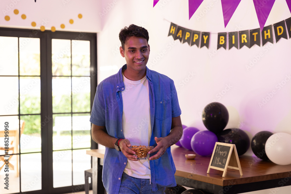 Fototapeta premium Indian teenage male holding bowl of snacks and smiling in party room, birthday banner, balloons