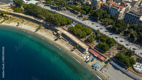 Fényképezés High-angle aerial view of a coastal promenade featuring a grey and white walkway, palm trees, and a urban beach alongside the crystal-clear and blue sea