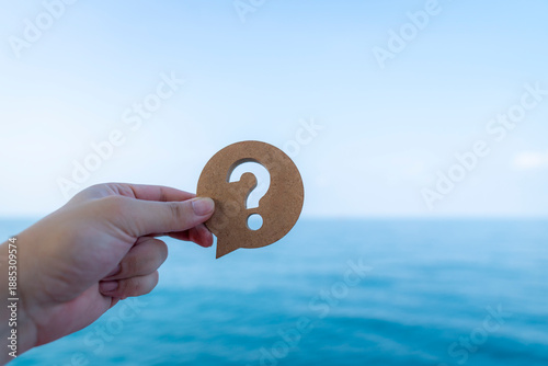 Fotografie Hand holding wooden house model over sea background, representing housing uncertainty, property risk, real estate decision making, unstable conditions, future planning, and changing environment