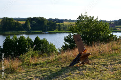 A close up on a wooden archery practice target with bow and arrows laying next to it located on a slippery slope covered with grass located next to a lake or pond seen in summer in Poland