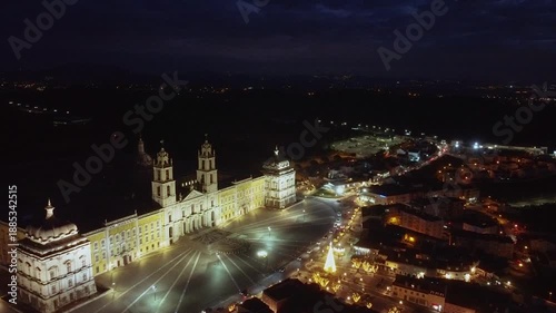 Cinematic aerial drone shot of Mafra National Palace at night, captured from an elevated lateral perspective, showcasing the illuminated facade and surrounding square under dark sky.