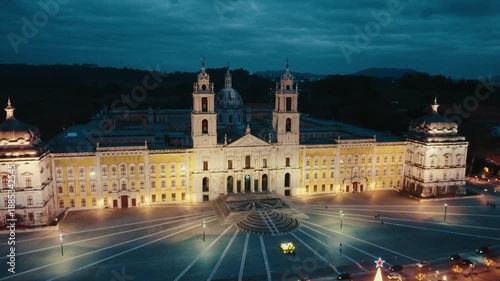 Aerial drone approach toward Mafra National Palace at dusk, moving forward and slightly descending, ending with close lateral perspective. Illuminated historic architecture for travel tourism use.