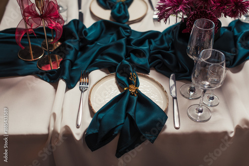Luxurious banquet table setting with beige tablecloth, green napkins in gold holders, red candles in golden candlesticks and ceramic plates. Refined festive dining decor.
