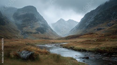 A stream flows through a valley in Glen Coe, Scotland. Mountains rise on either side under a cloudy, misty sky in the Scottish Highlands