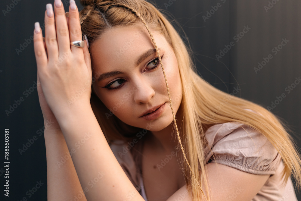 Fototapeta premium Young blonde woman with braids and a ring posing with her hands on her head and looking away from the camera, expressing a sense of contemplation or thoughtfulness against a dark background