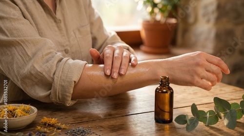 Woman applies essential oil to her arm from a brown glass bottle placed on a wooden table, surrounded by dried herbs and a potted plant, emphasizing a natural wellness atmosphere