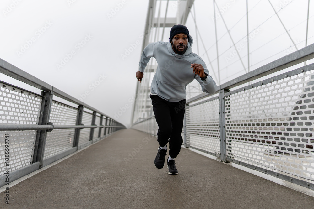 Naklejka premium Low-angle view of a Black male runner sprinting on a foggy urban bridge, holding a water bottle. Dynamic motion, cardio training, endurance and focus in modern outdoor fitness scene