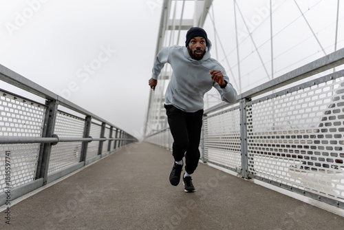 Billede på lærred Low-angle view of a Black male runner sprinting on a foggy urban bridge, holding a water bottle