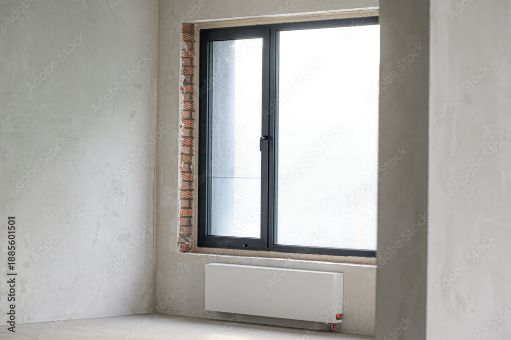 Fototapeta premium Bare room with unfinished walls; large black-framed window with brickwork visible on one side; white radiator mounted below window; minimalistic, empty space, natural light.