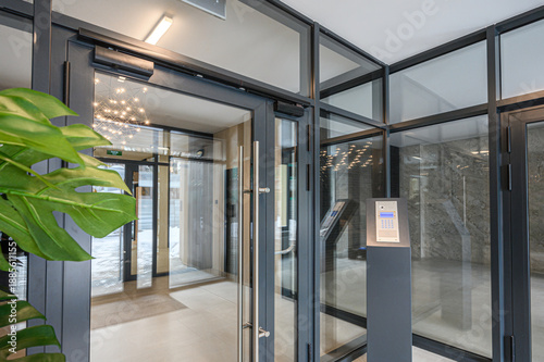 Modern entrance with glass doors, access control panel, sleek metal framing, indoor plants, polished floor, stylish lighting, clean minimalist design.