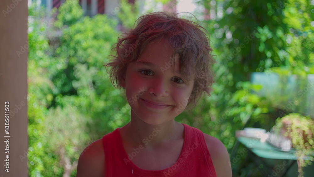 Fototapeta premium Smiling boy with tousled hair stands outside in vibrant green garden wearing red tank top enjoying carefree moment on summer day