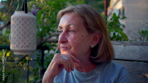 Senior woman gazes thoughtfully into distance while seated outdoors on wooden bench, surrounded by garden plants and soft evening light in tranquil setting