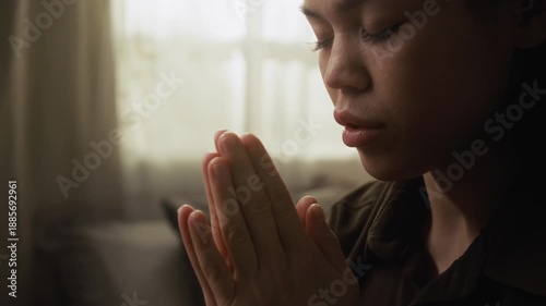 Close-up view of serene young woman with closed eyes and folded hands praying in morning with soft light filtering through window in background