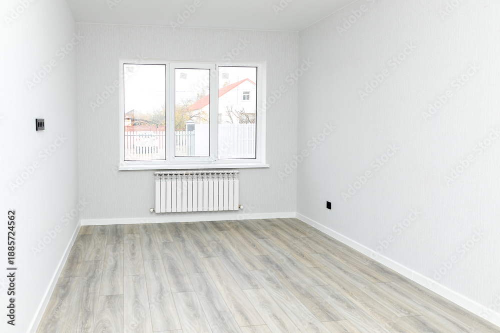 Obraz premium Empty interior room after renovations with white freshly painted walls and big window, allowing natural light, with a wooden floor, a radiator beneath the windows