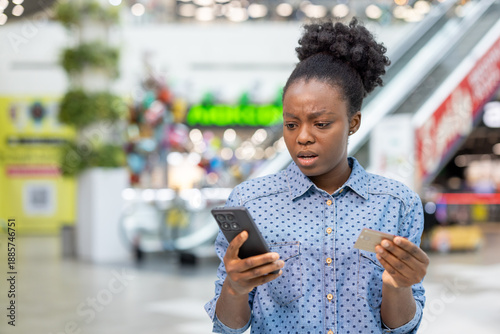 Bild auf Leinwand Shocked young woman in a mall holding credit card and smartphone, reacting to pa