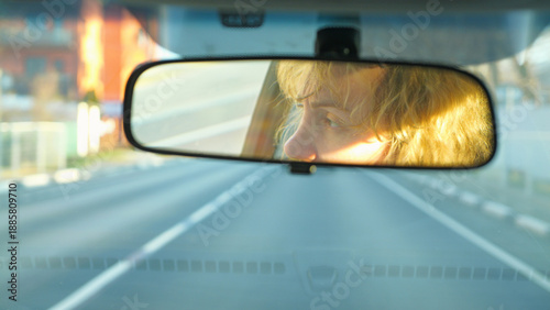 A woman's face reflects in a car's rearview mirror during a road trip. The scene, shot from a driver's POV, captures golden hour light, conveying a mood of travel and introspection